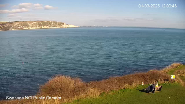 A scenic view of the ocean on a sunny day. The water is calm and reflects the blue sky, which has a few fluffy white clouds. In the foreground, there is a grassy area with some bushes. A couple of people are sitting on a bench, enjoying the view. In the distance, white cliffs can be seen along the shoreline, and there are small boats in the water. A sign is located near the bench. The image has a timestamp indicating it was taken on March 1, 2025, at noon.