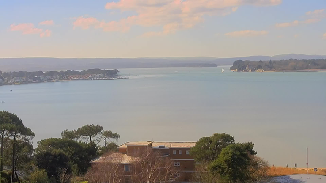 A serene waterfront scene featuring a calm, blue body of water. On the left side, there are small boats and a dock area, with houses lining the shore. Lush green trees are visible in the foreground, and a sandy area is present near the water's edge. In the distance, gentle hills rise and a few white sailboats can be seen sailing on the water. The sky is light blue with soft, fluffy clouds scattered throughout.