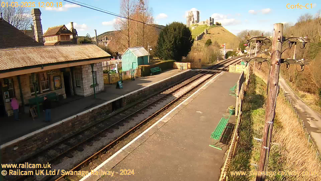 A railway station platform is shown with two sets of train tracks running parallel. Stone-built station buildings are on the left, featuring a pitched roof and large windows. A few people are standing on the platform. To the right, there are green benches and a small wooden shelter. In the background, a castle ruins atop a hill can be seen against a blue sky with scattered clouds. The scene is bright and sunny.