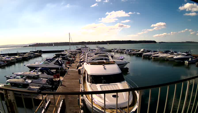 A marina scene featuring several boats docked in calm waters. The foreground shows a large white boat with a curved top. Surrounding it are numerous smaller boats of various colors, mostly white and blue, tied to the wooden pier. In the background, gentle waves reflect the sky, which is partly cloudy with a hint of sunset hues. Sailboats are visible further out on the water.