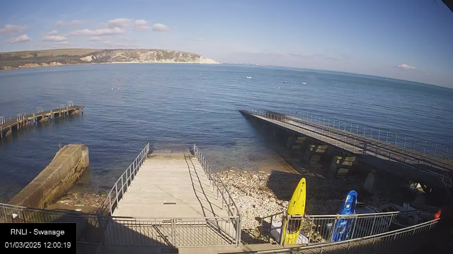 A view of a calm sea under a clear blue sky. The shoreline features a concrete ramp leading down to the water, surrounded by rocky ground. To the right, there are two kayaks, one yellow and one blue, stored upright on the shore. In the background, gentle hills rise with greenery, while a distant white cliff is visible along the coastline. A wooden pier extends into the water from the left side of the image.