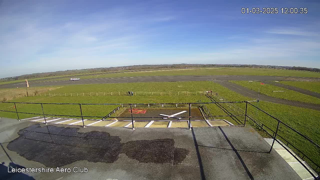 A clear blue sky stretches overhead, with a few wispy clouds. The image shows a grassy airport runway with a small white airplane preparing for takeoff in the background. In the foreground, there is a flat surface with a white cross marking and some wet spots, possibly from recent rain. Two figures, appearing to be people, stand on the runway near a wooden fence, observing the area. A windsock is visible to the left, indicating wind direction. The scene captures an expansive view of the airstrip and surrounding landscape.