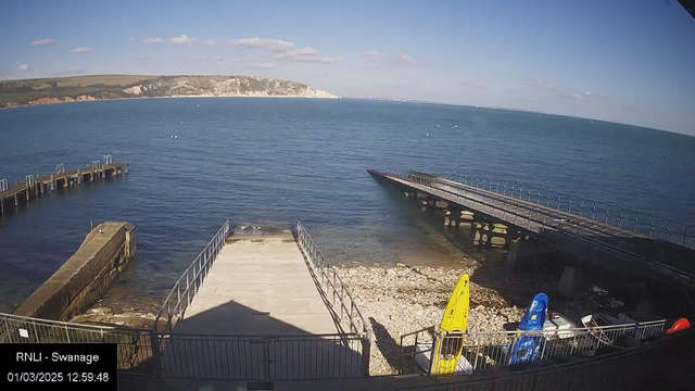 A view of a tranquil coastline featuring a clear blue sea under a bright sky with a few clouds. In the foreground, there are several boats, including a yellow and a blue kayak, positioned near a stony beach. Two piers extend into the water from the left side of the image, one with a railing and another that appears to lead to a boat ramp. The distant shore has white cliffs and green hills behind it, creating a picturesque backdrop.