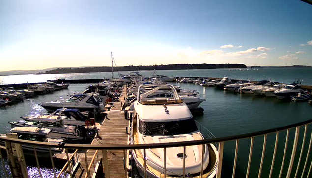 A scenic view of a marina under a clear blue sky. The image includes numerous boats docked at a wooden pier. A large white boat is prominent in the foreground, with smaller boats surrounding it. The water is shimmering in the sunlight, and there are green hills in the distance, along with a few clouds in the sky.