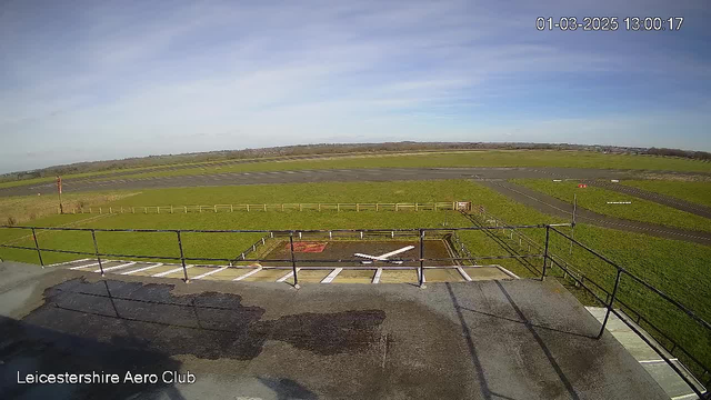 A wide view of a grassy airfield under a clear blue sky. In the foreground, there is a concrete area with white markings resembling a small aircraft. A wooden fence runs along the perimeter of the field. In the background, the airfield features a black runway with yellow markings. The scene appears peaceful and open, with no obstacles in sight. The timestamp in the upper right corner indicates the image was taken on March 1, 2025, at 1:00 PM.