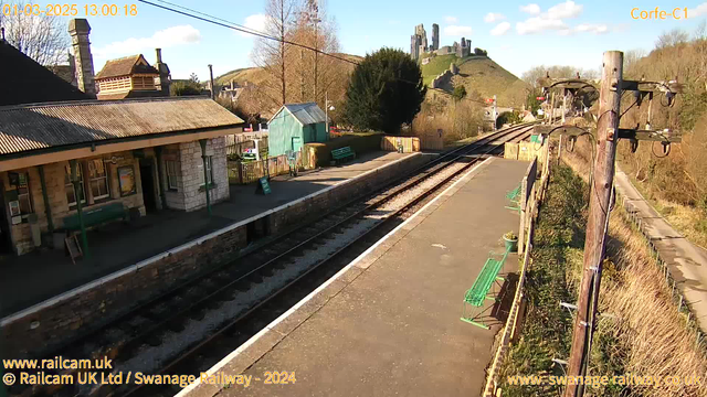 A serene railway station scene with stone buildings and benches. The platform is empty, and two railway tracks stretch into the distance. A small green shed is positioned near the station. In the background, a large hill rises featuring a castle, surrounded by trees and a clear blue sky dotted with a few clouds. The station includes signs and a fence along the path, under a bright, sunny atmosphere.