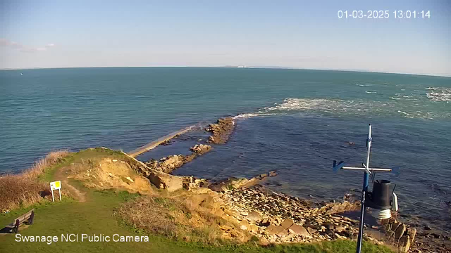 A coastal scene showing the ocean with gentle waves, rocks along the shoreline, and a grassy area in the foreground. There is a bench and a sign on the left side. A weather station structure with blue and black components stands on the right. The sky is mostly clear, and the water appears calm and blue.