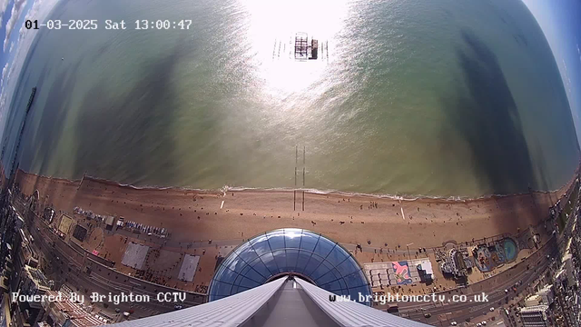 Aerial view of a beach with golden sand and gentle waves. The sun reflects brightly on the water, creating a shimmering effect. People walk along the shoreline, and some are visible on the sand. In the distance, a pier extends into the sea. The foreground shows a circular glass structure, possibly part of a viewing platform, with a metallic tower reaching upward. The sky is partly cloudy, with patches of blue visible. The date and time stamp is displayed in the upper left corner, reading "01-03-2025 Sat 13:00:47."