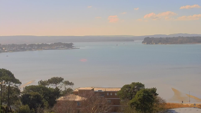 A wide view of a calm water body with a gentle slope of land on the left side featuring trees and buildings. In the distance, there are several boats and a small marina with docks along the shoreline. The background shows a horizon of gentle hills under a clear sky with a few wispy clouds. The sunlight creates a soft glow on the water's surface, reflecting shades of blue and hints of golden hues near the shoreline. The scene conveys a serene and peaceful atmosphere.