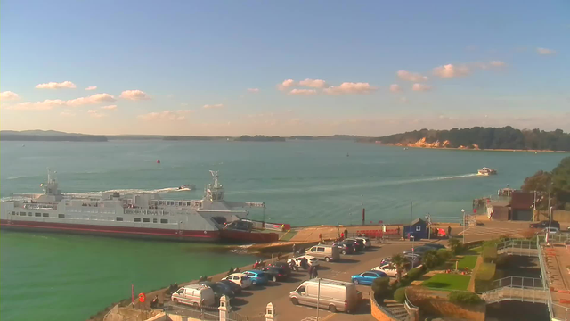 A view of a waterfront scene featuring a ferry docked at a pier on the left side. In the foreground, there is a parking area with several cars, including white and blue vehicles, and a few people walking. The water is calm and turquoise, with a few boats in the distance. A green hillside and cliffs are visible in the background under a clear blue sky with scattered clouds.