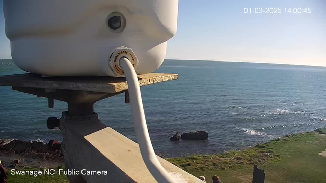 A white container sits atop a sturdy surface, with a tube extending from its base. In the background, the calm sea reflects sunlight, and the shoreline is visible with some rocks and a grassy area. The scene is clear and sunny, evoking a peaceful coastal atmosphere.