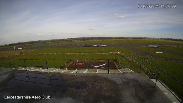 A view from a webcam showing the Leicestershire Aero Club. In the foreground, there is a flat rooftop with a railing, and a white runway marking on the ground. The background features a grassy airfield with two small aircraft on the runway. The sky is mostly clear with a few clouds. The image timestamp shows it is 2:00 PM on March 1, 2025.