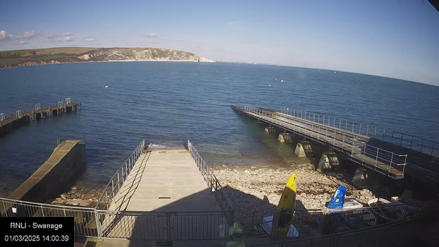 A clear blue sea stretches out under a bright sky with a few clouds. There are two piers extending into the water, one with a wooden deck and the other made of metal. To the right, there are yellow and blue kayaks resting on the shore, with rocky beach areas partially exposed. In the distance, cliffs rise along the coastline. The scene is peaceful and captures a sunny day by the water.