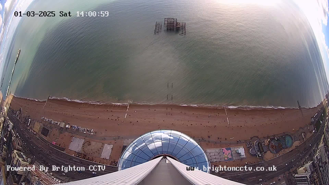 An aerial view from a tall structure overlooking a beach and the sea. The beach has a sandy area with small dots that likely represent people. There is a pier extending into the water on the left side of the image. In the sea, there are remnants of a dock or structure. The foreground shows part of the tall building with a blue dome, and there are colorful patterns on the ground near the base of the building. The sky is partially cloudy, and the time and date are displayed at the top of the image.