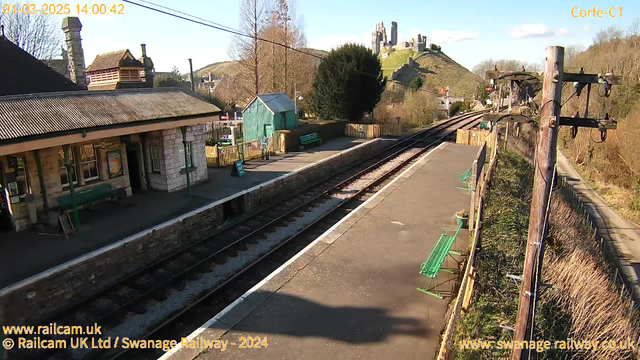 A railway station scene captured from a webcam on a clear day. The foreground shows a platform with several green benches, bordered by a stone wall. On the left, there is a building with a sloped roof, partially obscured by trees. In the background, a hill rises with a castle visible at the top, surrounded by greenery. The railway tracks run parallel along the platform, leading towards the horizon. The sky is blue with a few clouds.