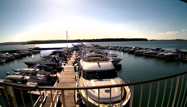 A view of a marina on a sunny day, with multiple boats docked at a wooden pier. The water is calm and reflects the sunlight. In the background, there is a green shoreline and a clear blue sky with a few clouds. Several boats of various sizes are moored along the dock, with some visible in the distance. The scene conveys a peaceful and recreational atmosphere by the water.