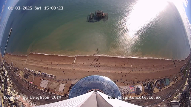 A high-angle view of a sandy beach with scattered people walking along the shore. The beach is bordered by a promenade and seaside structures, including a large circular building with a glass dome in the foreground. The sea is calm, with gentle waves and a reflection of sunlight on the water. In the distance, a pier extends into the ocean, while a sunken structure is visible offshore. The time and date are displayed at the top of the image.