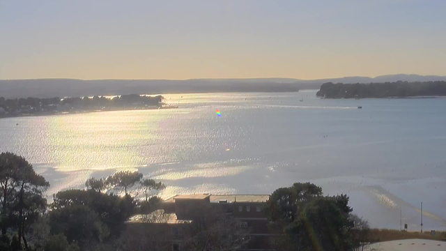 A scenic view of a calm body of water reflecting sunlight, creating a shimmering effect. The horizon is lined with trees and distant hills. Small boats can be seen floating on the water, and faint outlines of structures are visible along the shoreline. The sky is clear with a soft gradient as the day transitions towards evening, enhancing the tranquil atmosphere.