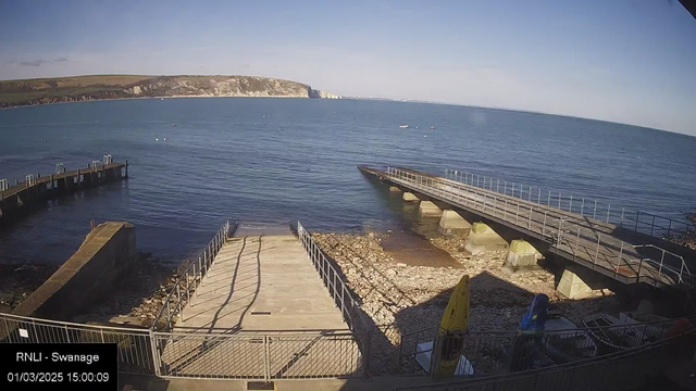 A coastal scene showing a calm blue sea under a clear sky. On the left, there is a wooden jetty extending into the water with several boats visible. In the foreground, a concrete ramp leads down to the shoreline, which is rocky and scattered with small stones. To the right, there are two kayaks resting on the ground, one yellow and the other blue. In the background, a white cliff is visible along the coastline.