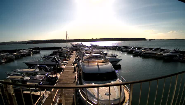 A marina scene with numerous boats moored in calm water. The sun is low in the sky, creating a shimmering reflection on the water's surface. A wooden dock leads to the boats, while a railing is visible in the foreground. The background features hills and a clear sky.