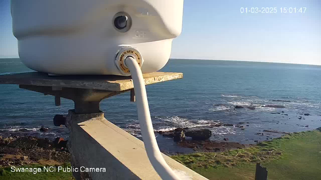 A white container sits on a platform overlooking a coastal scene. A hose extends from the container towards the edge. Below, the rocky shoreline is visible, with waves gently lapping against the rocks. In the background, a clear blue sky meets the sea, creating a serene atmosphere. A timestamp in the corner indicates the date and time of the image.