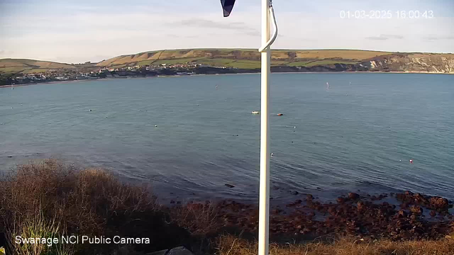 A coastal view showing calm water with gentle waves. In the foreground, there are some rocky areas and patches of grass. A white flagpole is positioned on the right side of the image. In the background, green hills rise above a small town along the coastline, with a few boats visible on the water. The sky is mostly clear with some clouds, indicating a pleasant day.