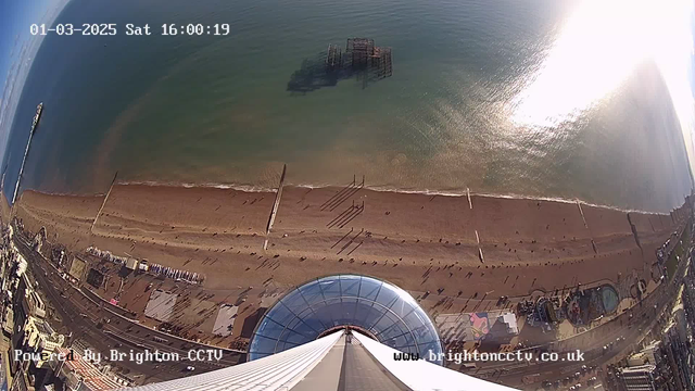 A panoramic view from a tall structure overlooking a beach. The beach features a long stretch of sandy shoreline with small waves lapping at the edges. Shadows cast by people walking along the beach create patterns in the sand. A pier extends out into the sea on the left side, and remnants of an old structure can be seen partially submerged in the water. To the right, there are buildings and attractions near the beach, with a clear blue sky above and sunlight reflecting off the water. The date and time are displayed in the top left corner.