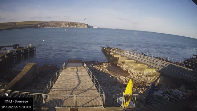 A view of a coastal area featuring a calm sea under a clear blue sky. In the foreground, there is a wooden pier extending over the water, with a ramp leading down from it. To the left, a rocky area is visible along the shoreline, and a few boats are moored nearby. On the right side, there are multiple boats, one yellow kayak, and some equipment on the shore. The date and time displayed at the bottom indicate it is March 1, 2025, at 16:00.