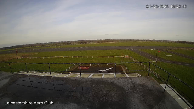 A view from a webcam overlooking an airfield at Leicestershire Aero Club. The scene includes a large green grassy area with a runway made of asphalt. There are low wooden fences lining the sides of the runway. In the foreground, a white X is painted on a dark patch of ground, likely indicating a designated area. The sky appears mostly clear with light clouds. The time stamp on the image shows March 1, 2025, at 16:00.