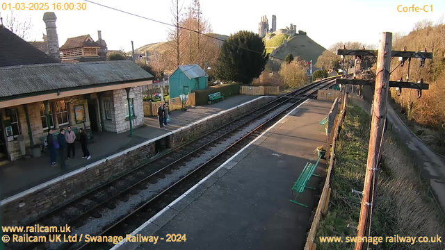 The image shows a railway station platform with several people standing near the edge. In the background, there is a historic castle atop a hill. The sky is clear with warm sunlight illuminating the scene. On one side of the platform, there is a building with a sloped roof and windows, while opposite are green benches and a wooden railing. Railway tracks run through the center of the image, leading into the distance. Trees and a small green shed are visible at the end of the platform.