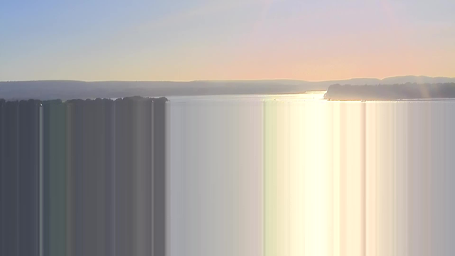 A scenic view of a body of water stretching to the horizon, with a soft gradient of colors ranging from pale blue to warm orange as the sun sets. The surface of the water reflects the sunlight, creating a shimmering effect. In the distance, faint outlines of hills and trees are visible along the shoreline. The sky is clear with no clouds, indicating a calm atmosphere.