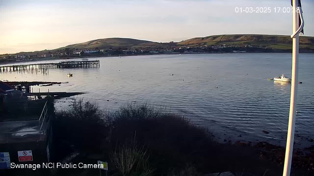 A view of a calm sea at sunset, with gentle waves rippling across the surface. In the foreground, a pier extends into the water, partially constructed with wooden pillars. A small white boat is anchored in the water, and several buoys float nearby. In the background, rolling green hills rise gently, dotted with houses. The scene is peaceful, with soft colors in the sky transitioning from light blue to pale orange.