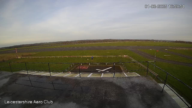 A view from a webcam situated on a terrace overlooking an airfield. In the foreground, there is a large white X on the ground, possibly indicating a landing area. A person wearing a blue jacket is standing on the grass near a small dog, with a wooden fence in the background. The airfield is primarily green with a few patches of gray asphalt and a windsock visible in the distance, indicating wind direction. The sky is mostly cloudy, and the scene appears calm and open. The name "Leicestershire Aero Club" is displayed at the bottom of the image.