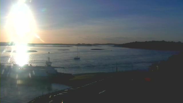 A sun setting over a calm body of water, casting bright reflections on the surface. In the foreground, there is a boat docked near the shore, with another sailing boat further out in the water. The background features gentle hills and islands on the horizon, while the sky transitions from blue to soft orange and pink hues. Shadows from nearby structures can be seen in the lower part of the image.