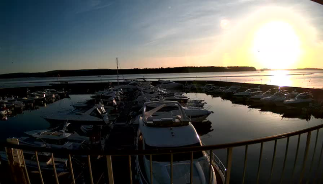 A scenic view of a marina at sunset, featuring multiple boats docked in calm water. The sun is low on the horizon, casting a golden glow across the sky and reflecting on the water's surface. Gentle ripples distort the reflection, while the silhouette of a distant shoreline is visible. A railing is partially visible in the foreground, framing the scene.