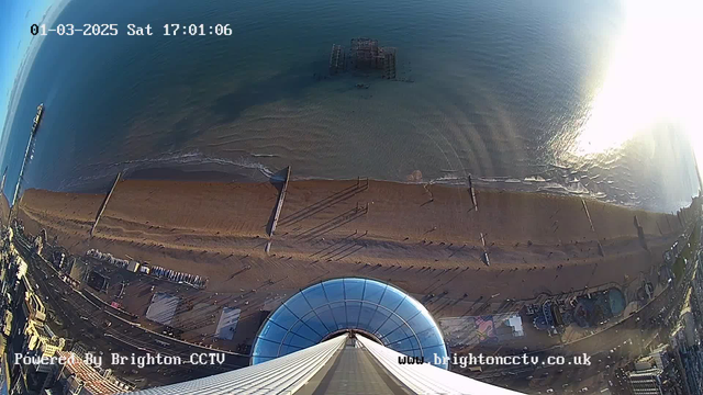 A bird's-eye view of a beach with a shoreline stretching across the lower part of the image. The image shows a calm sea with gentle waves, reflecting sunlight. At the far left, a pier extends into the water. In the distance, a structure resembling a ruined pier is partially submerged. The foreground features a large, circular glass structure, possibly an observation deck, with vertical lines stretching upwards. The surrounding area includes sandy beach, with a few small figures and beach furniture visible. The upper part of the image is clear blue sky, and the date and time are displayed in the top left corner.