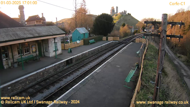 A railway station platform is visible, featuring a stone building with green benches and large windows. On the left, the roof has a slight slope, and a chimney is visible. In the background, a hill rises with ruins at the top, likely a castle, while trees frame the scene. The tracks are lined with gravel and lead off into the distance. The sky is clear, indicating sunset with warm lighting. A sign on the platform reads "WAY OUT."