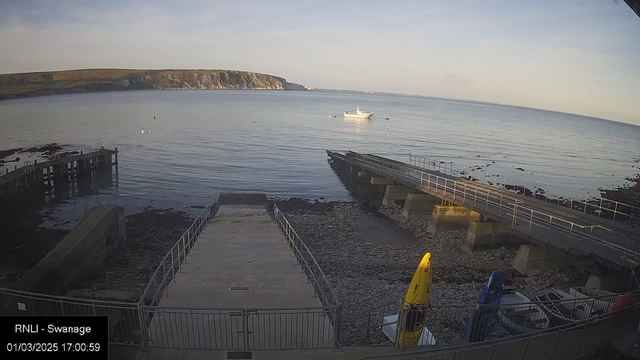 A calm seaside view shows a rocky shoreline with a boat dock on the left. The water is smooth and reflects the sky, with a boat floating in the distance. A long ramp extends from the dock into the water, and several small boats are tied up at the dock. In the foreground, colorful kayaks are resting on the ground near the dock. The background features a steep, grassy cliff rising in the distance under a clear sky. The overall scene is tranquil and scenic, evoking a serene coastal atmosphere.