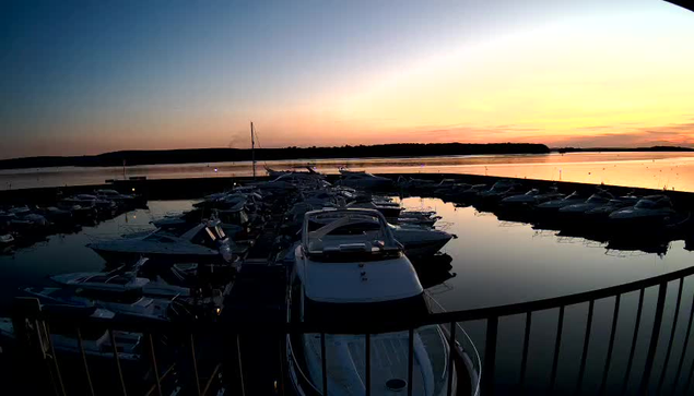 A view of a marina during sunset, with numerous boats docked in the water. The sky is painted in shades of orange and blue, reflecting on the calm surface of the water. In the foreground, there is a railing framing the scene. The background features a tree-lined shore against the horizon.