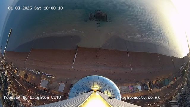 Aerial view of a sandy beach meeting the ocean, with a pier extending into the water. The scene is partially illuminated by the setting sun, casting soft light over the waves. In the foreground, there is a circular platform with a roof, suggesting a high viewpoint. People can be seen on the beach and along the promenade, while various attractions and buildings line the shore. The time and date displayed at the top show "01-03-2025 Sat 18:00:10."