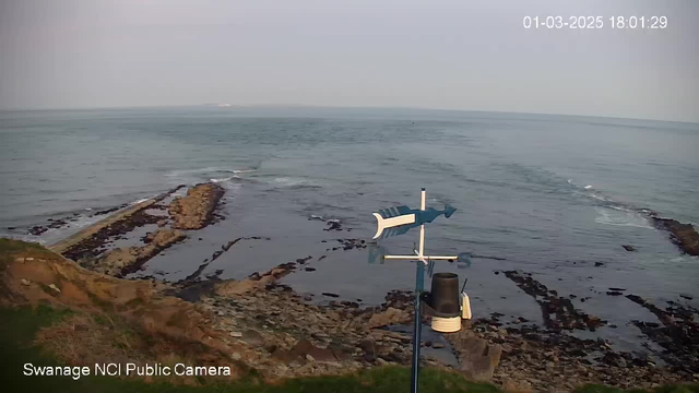 A coastal scene with a rocky shoreline extending into the sea. In the foreground, there is a weather vane shaped like a fish on a pole. The background shows calm water with gentle waves, and a soft, cloudy sky above. The image has a timestamp displaying the date and time at the top right corner.