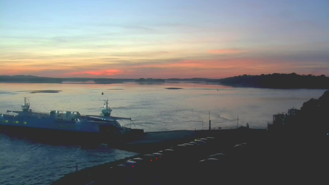 A waterfront scene at dusk featuring a large boat docked at a pier. The sky displays a gradient of colors, shifting from pale blue to soft pink and orange. The water reflects these colors and has gentle ripples. In the foreground, there are several parked cars lining the edge of the pier. On the right side, hints of a dark landscape and buildings are visible, while in the distance, faint outlines of hills can be seen on the horizon.