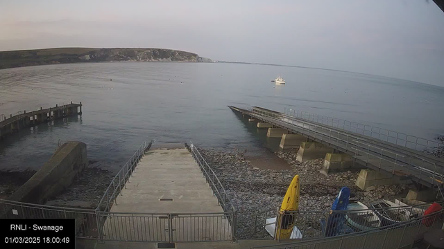 A calm seaside view shows a rocky shoreline with a boat ramp leading down to the water. To the right, a small white boat is anchored in the sea. Several kayaks in bright colors, including yellow and blue, are docked on the shore. In the background, a cliff rises above the water, partially covered by greenery. The sky is pale with soft clouds, indicating early evening light.