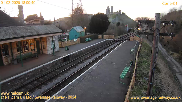 A railway station platform during early evening. The scene shows several green benches along the platform, with a stone wall running alongside the railway tracks. In the background, a hill is visible with ruins of a castle on top, surrounded by trees. The sky is light, indicating sunset, and there are power poles with wires running overhead. A sign on a fence reads "WAY OUT." On the left side, there's a building with a sloped roof and large windows, likely a station building. The atmosphere is calm and quiet.