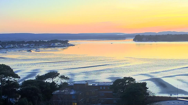 A calm body of water reflects warm tones of orange, pink, and blue from the sky during sunrise or sunset. In the foreground, trees and buildings are visible, with some boats scattered across the water. The horizon features distant hills silhouetted against the colorful sky. Gentle ripples create patterns on the water's surface.