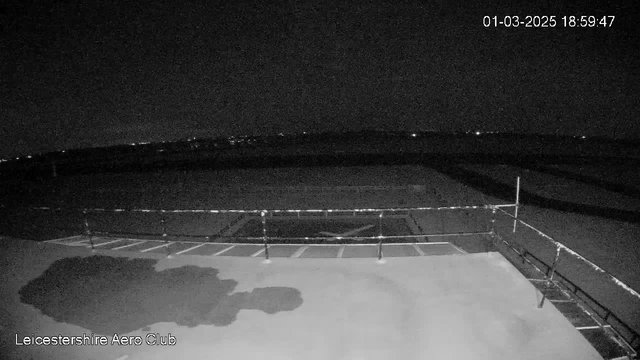 A dark, nighttime view from a webcam at the Leicestershire Aero Club. The foreground features a fenced area with a flat surface and faint outlines of what appears to be parking spaces. In the background, there are distant lights visible on the horizon, suggesting the presence of a landscape or cityscape. The image has a low level of illumination. The timestamp indicates the date and time as March 1, 2025, at 18:59:47.