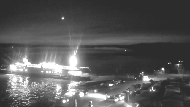 A black and white image of a harbor at night. A bright ferry is docked on the left side of the image, illuminated by several lights. The water is reflective, showing the glow from the ferry. On the right, there are parked vehicles along the waterfront, and a few scattered lights suggest nearby buildings. The sky is dark with a faint light visible in the upper right corner, possibly a star or distant object.