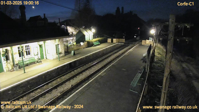 A nighttime image of an empty railway station platform. On the left, there is a building with large windows and green accents, illuminated by yellow light. Several green benches are positioned along the platform. A wooden fence runs alongside the platform, with a sign indicating "WAY OUT." The railway tracks are visible, leading away from the platform. In the background, the silhouette of buildings is seen against the dimly lit sky, with a crescent moon visible in the upper left corner.