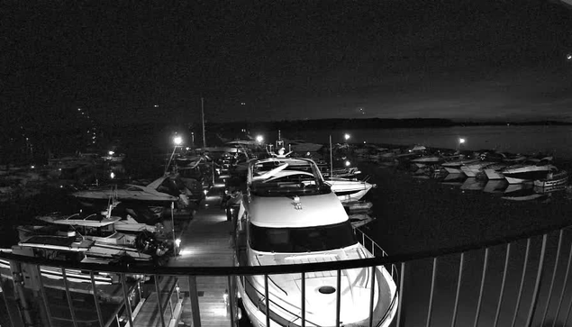 A nighttime marina scene featuring several boats docked along a wooden pier. The image is predominantly black and white, with some illuminated areas showing lights from the boats and reflections on the water. The sky is dark with a faint horizon line.