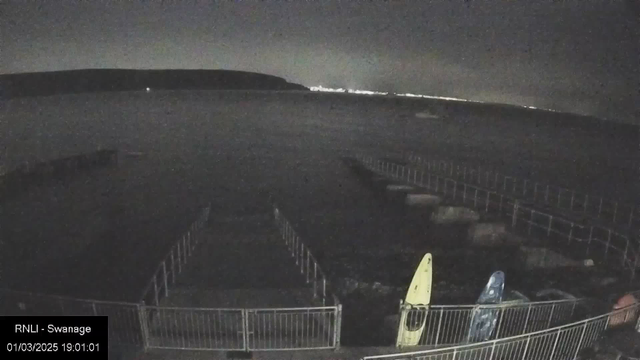 A dark scene of a waterfront at night. In the foreground, there are several brightly colored kayaks, one yellow and one blue, resting against a railing. Beyond the railing, a pier extends into the water, which appears calm. In the distance, faint lights from a coastal town can be seen along the horizon, and a low hill or cliff outlines the background. The sky is dark with hints of light. The time stamp indicates it is evening.